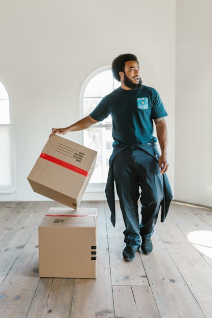 A young woman with long, curly hair and a neutral expression is standing inside a home, holding two large cardboard boxes with taped seams and handles, one labeled with the handwritten word 'toys.' Sh